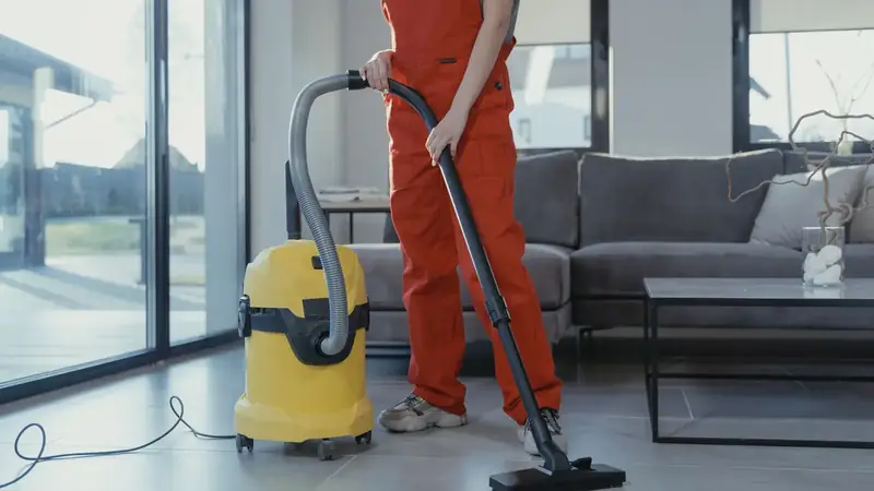 Reinigungskraft in Arbeitskleidung saugt den Boden eines Wohnraums